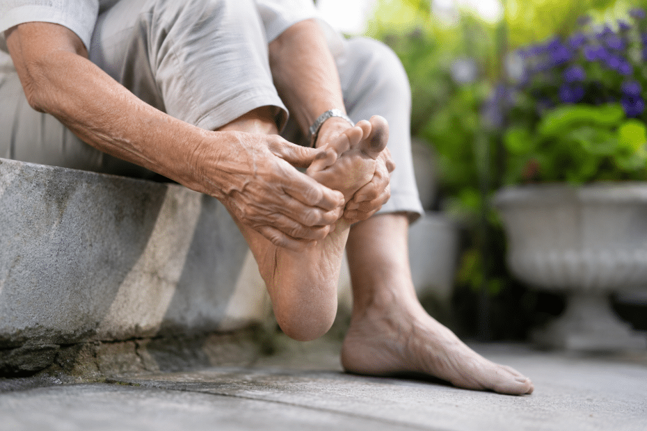 Elderly person sits on a stone step, gently holding and examining their bare foot outdoors in a garden.